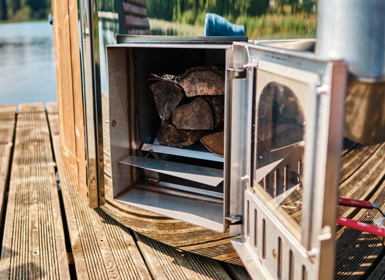 Geöffneter Metallofen eines Feuerbad Hot Tubs SOPHIA mit eingelegtem Brennholz, sichtbar auf einem Holzsteg am See