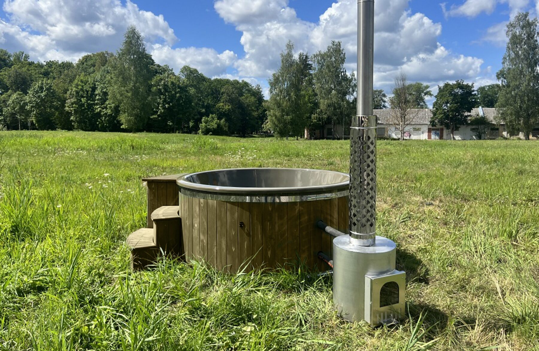 Feuerbad Hot Tub DIANA steht auf einer grünen Wiese mit Holzverkleidung, einem externen Metallofen mit Schornstein sowie einer Holz-Einstiegstreppe, umgeben von Bäumen und bewölktem Himmel