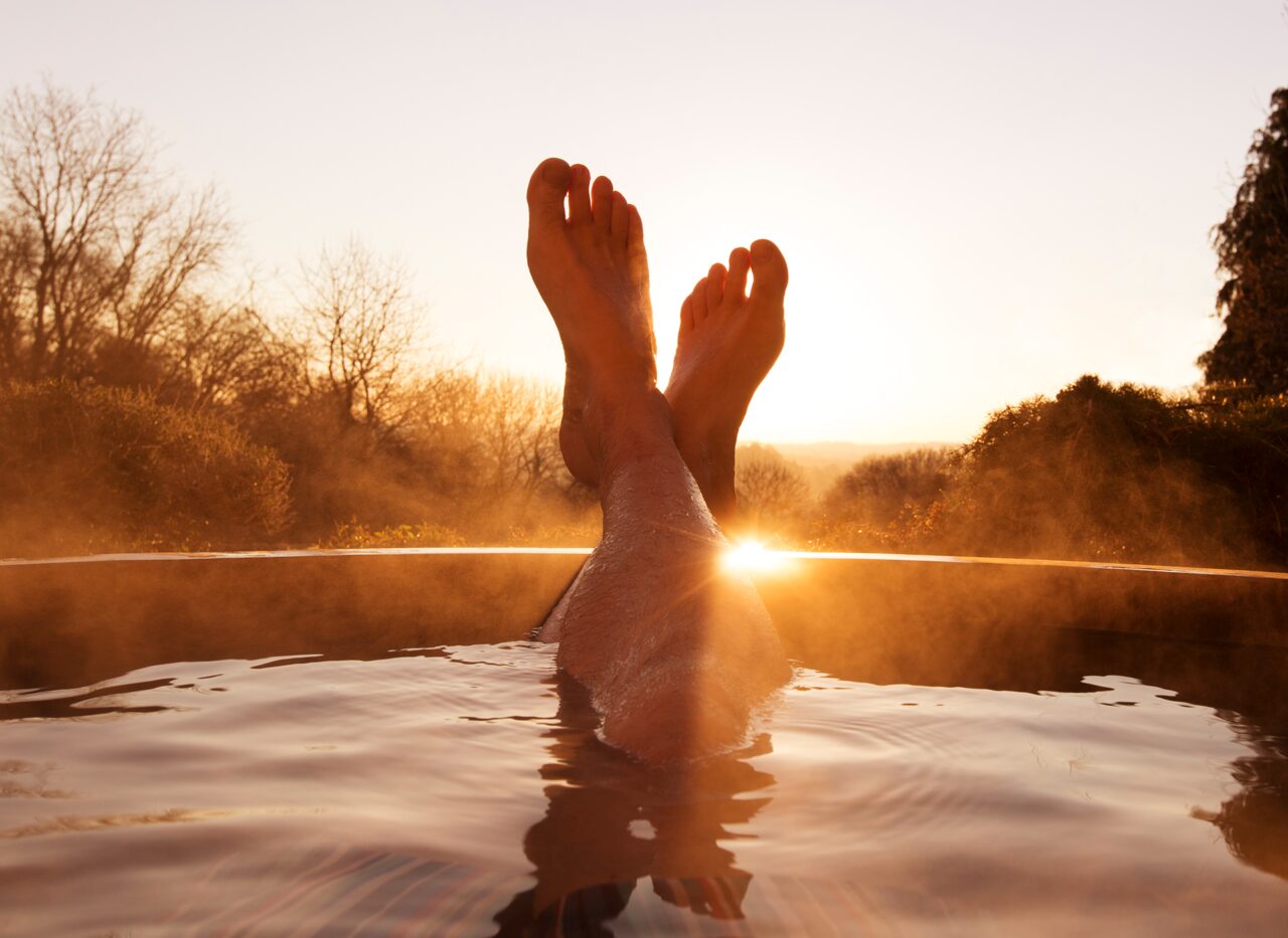 Eine Person entspannt im Wasser bei Sonnenuntergang, mit angehobenen Beinen und einer Naturlandschaft im Hintergrund