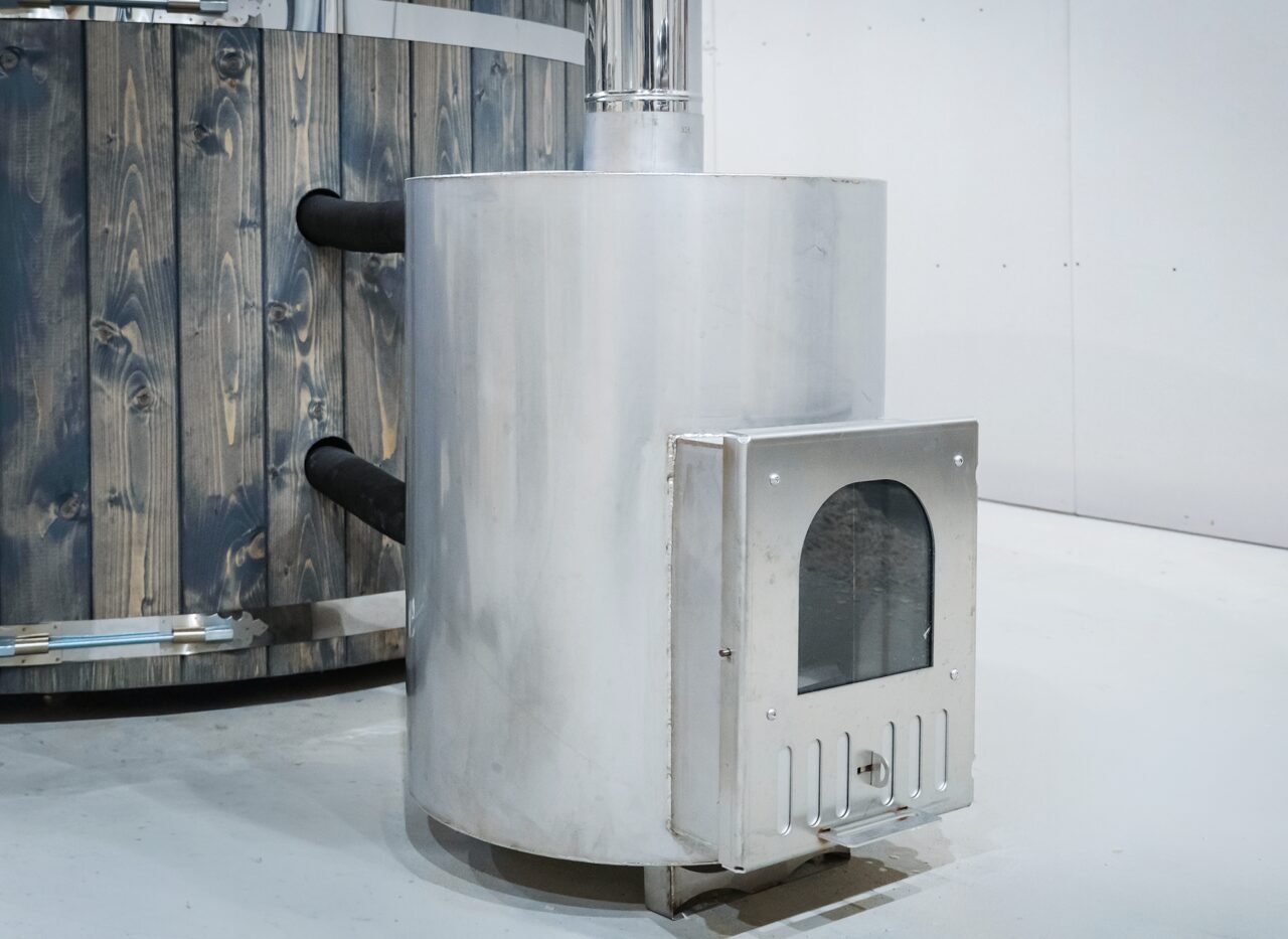 Detailansicht des externen Metallofens des Feuerbad Hot Tubs DIANA, mit Feuerraumtür, Anschlussleitungen zum Badefass und robuster Konstruktion neben der Holzverkleidung