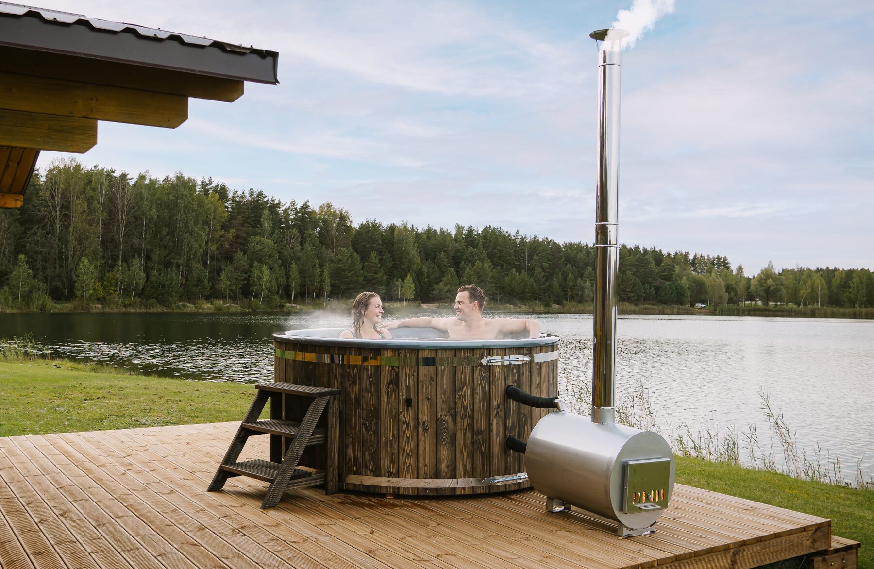 Feuerbad Hot Tub LUNA aus Holz auf einem Holzdeck, mit externem Metallofen und Schornstein, Holz-Einstiegstreppe und grüner Naturlandschaft im Hintergrund. Zwei Personen entspannen sich im dampfenden Hot Tub.