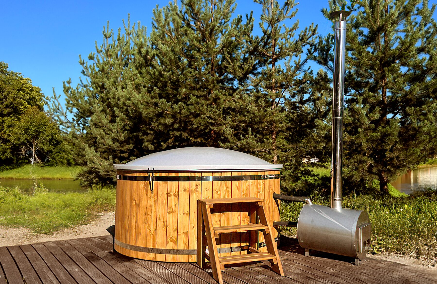 Feuerbad Hot Tub LUNA aus Holz mit geschlossener Abdeckung auf einem Holzdeck, mit externem Metallofen und Schornstein, Holz-Einstiegstreppe und grüner Naturlandschaft im Hintergrund
