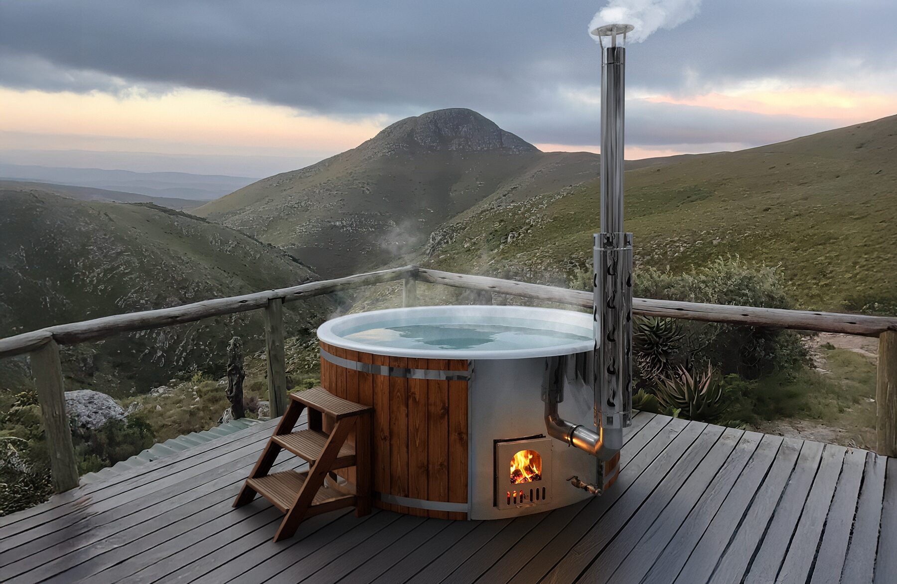 Hot Tub SOPHIA von Feuerbad aus Holz auf einer Terrasse mit Holzgeländer und Blick auf eine grüne Berglandschaft bei bewölktem Himmel