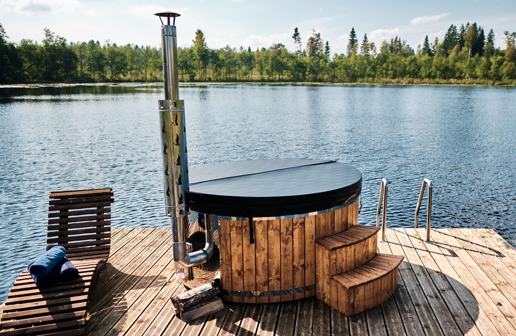 Feuerbad Hot Tub SOPHIA mit schwarzer Abdeckung auf einem Holzsteg am See mit Metallofen und Schornstein, Einstiegstreppe und Liegestuhl, umgeben von Wald