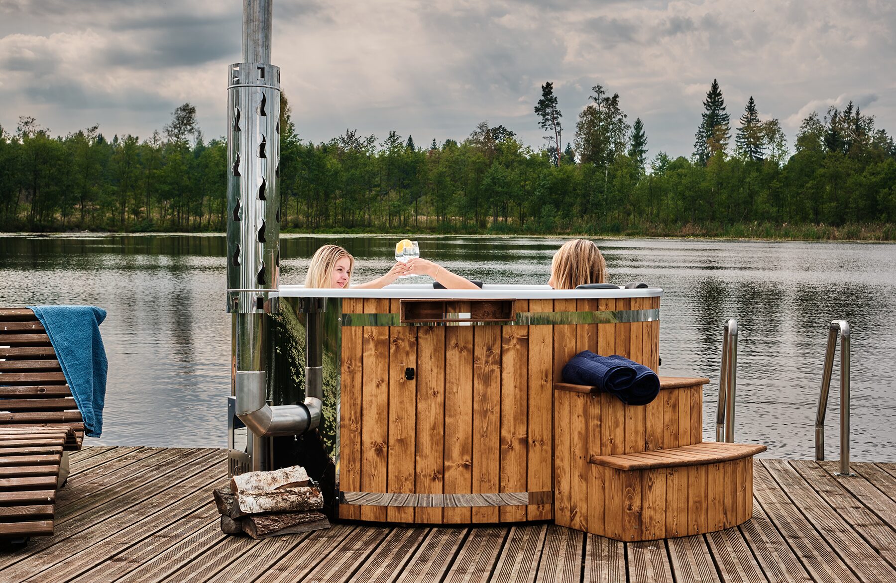 Feuerbad Hot Tub SOPHIA, in dem zwei Personen entspannen, steht am See mit Metallofen und Schornstein, Einstiegstreppe und Handtüchern, umgeben von Wald