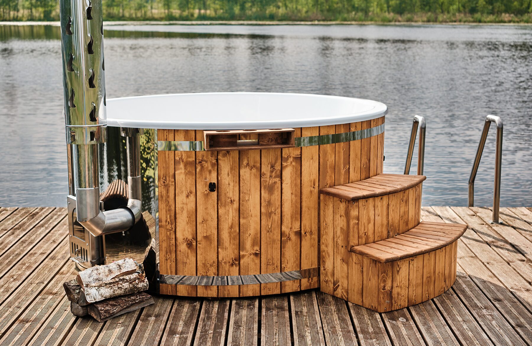 Feuerbad Hot Tub SOPHIA mit weißer Innenwanne auf einem Holzsteg am See, mit Metallofen und Schornstein, Holztreppe zum Einstieg und Brennholz daneben