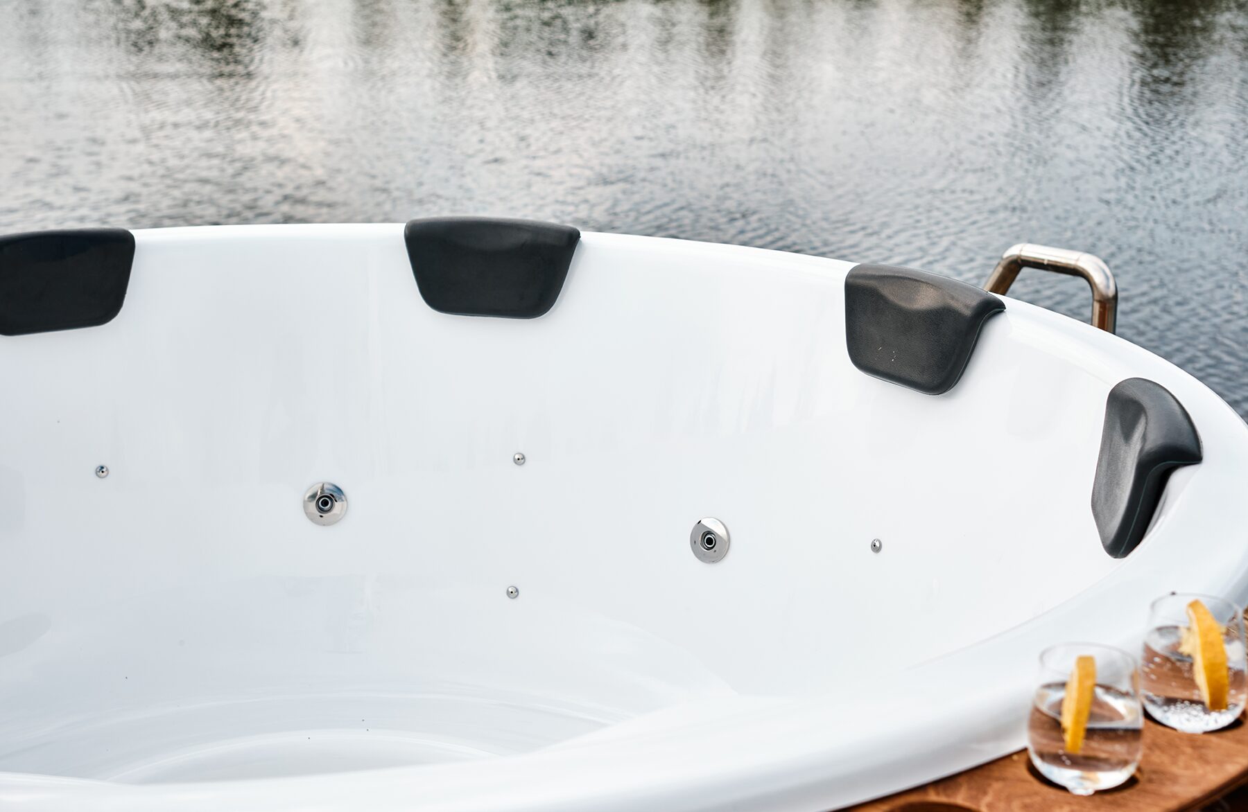 Detailansicht Feuerbad Hot Tub SOPHIA mit einer weißen Innenwanne mit integrierten Düsen und schwarze Nackenstützen mit einem See im Hintergrund und Getränken am Rand