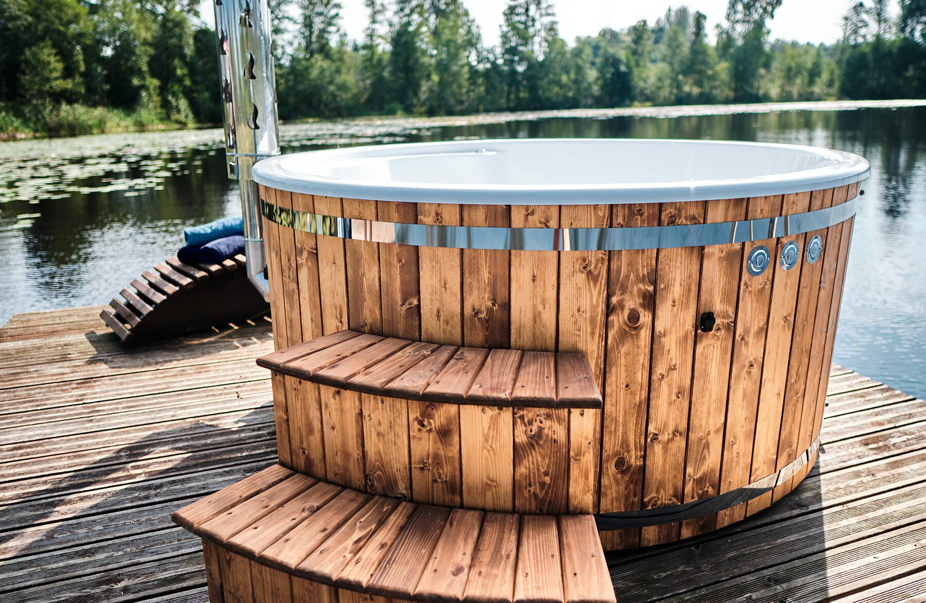 Feuerbad Hot Tub SOPHIA mit einer weißen Innenwanne und integrierten Düsen auf einem Holzsteg am See, mit Holz-Einstiegstreppe, Edelstahlband und Waldlandschaft im Hintergrund