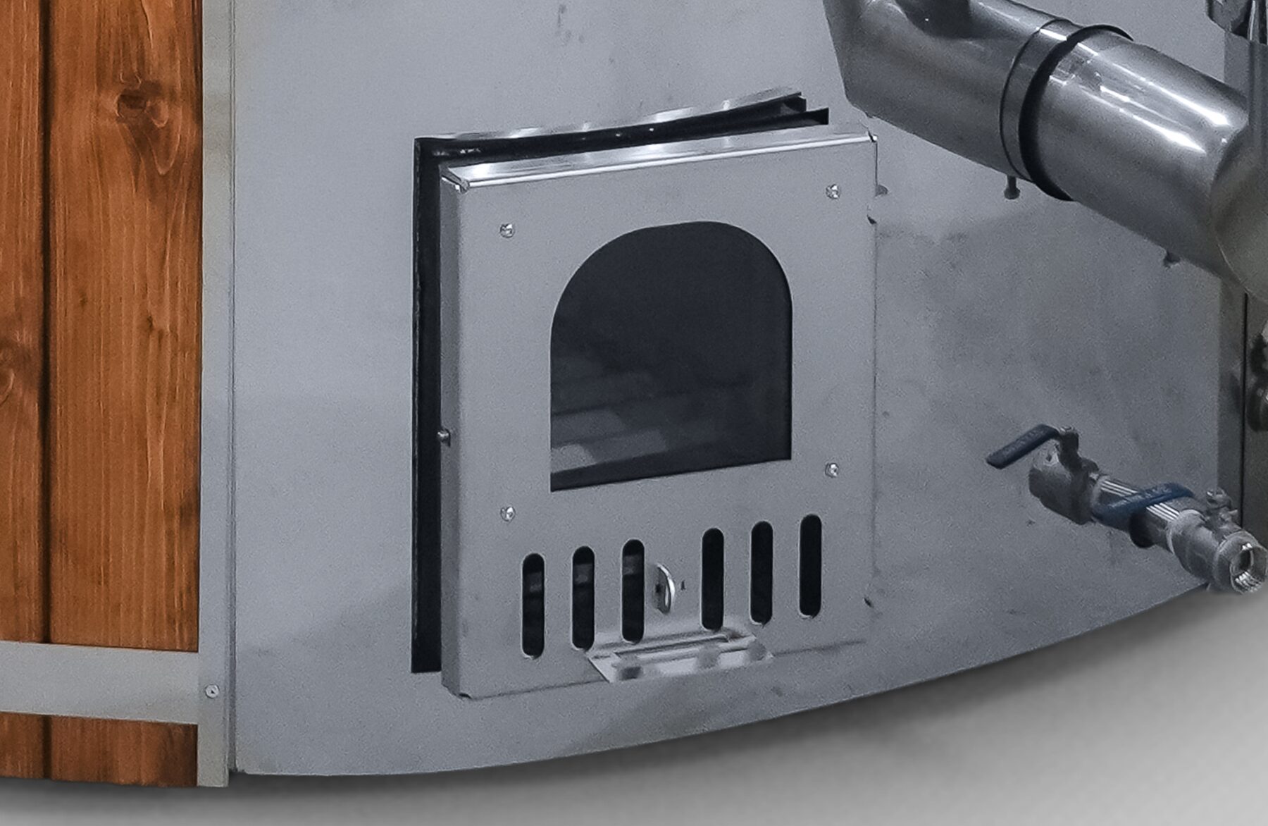 Detailansicht des Metallofens eines Feuerbad Hot Tubs SOPHIA mit Feuerraumtür, Luftschlitzen, Rohranschluss und Wasserablassventil an der Außenseite der Wanne