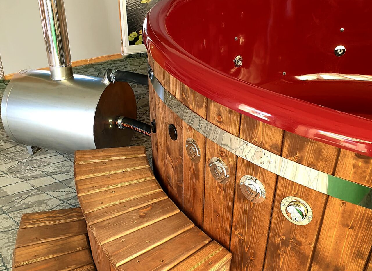 Detailansicht eines Feuerbad Hot Tubs mit roter Innenwanne, integrierten Massagedüsen und Holzverkleidung, ergänzt durch eine Holz-Einstiegstreppe und einen angeschlossenen Edelstahl-Holzofen im Innenbereich