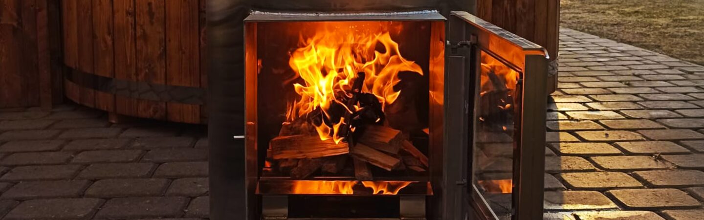 Zu sehen ist ein Metallofen mit geöffneter Tür, in dem ein Feuer über aufgeschichteten Holzscheiten brennt. Der Ofen steht auf einem gepflasterten Außenbereich vor einem Hot Tub.
