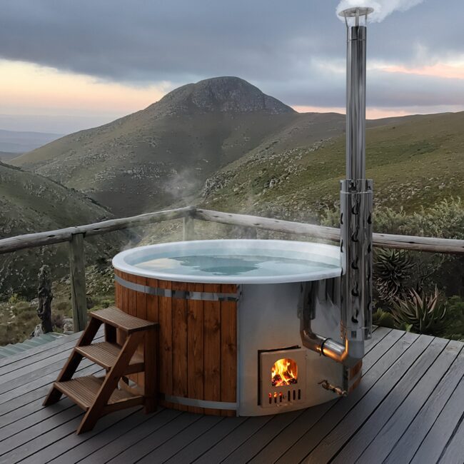 Hot Tub SOPHIA von Feuerbad aus Holz auf einer Terrasse mit Holzgeländer und Blick auf eine grüne Berglandschaft bei bewölktem Himmel