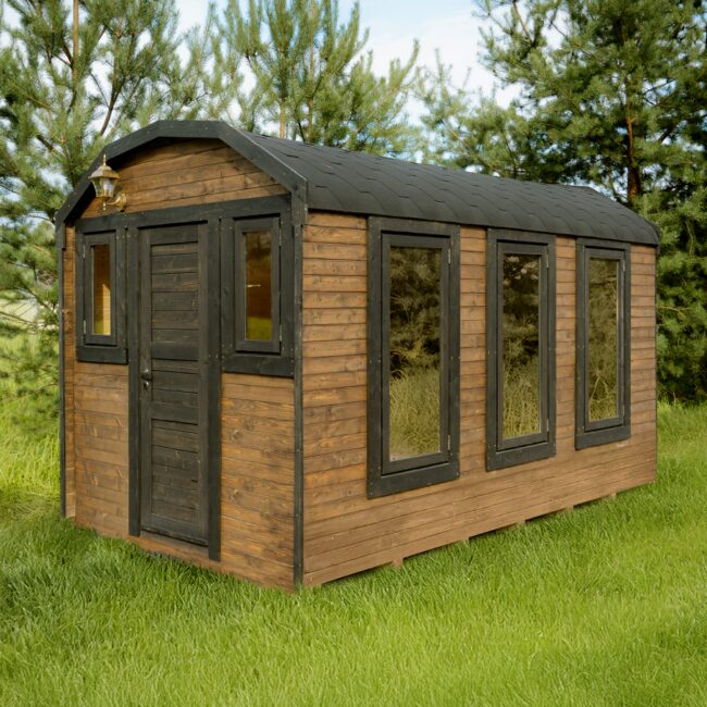 Feuerbad Sauna CERES auf einer Wiese, umgeben von Büschen und Bäumen. Im Hintergrund sieht man einen See und blauen Himmel