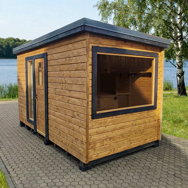 Außenansicht eines Feuerbad Tiny House DELUXE mit Holzverkleidung und dunklen Fensterrahmen, auf gepflastertem Untergrund direkt am See, umgeben von grüner Wiese, Bäumen und ruhiger Naturlandschaft