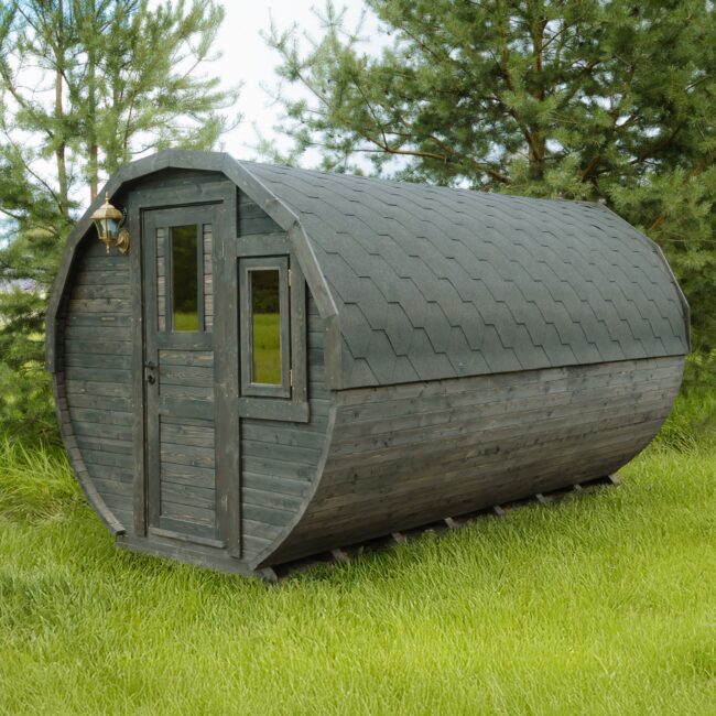 Außenansicht des Feuerbad Tiny House JUNO mit dunkler Holzverkleidung und gewölbter Form, ausgestattet mit Eingangstür, Seitenfenster und Dachschindeln, platziert auf einer grünen Wiese und umgeben von Bäumen