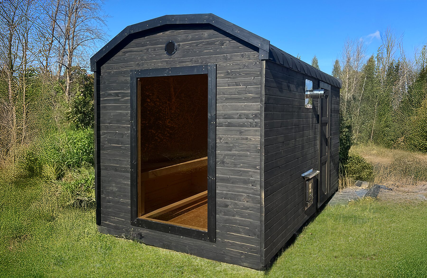 Feuerbad Sauna CERES mit dunkler Holzverkleidung und Glastür steht auf einer Wiese, umgeben von Büschen und Bäumen, unter blauem Himmel
