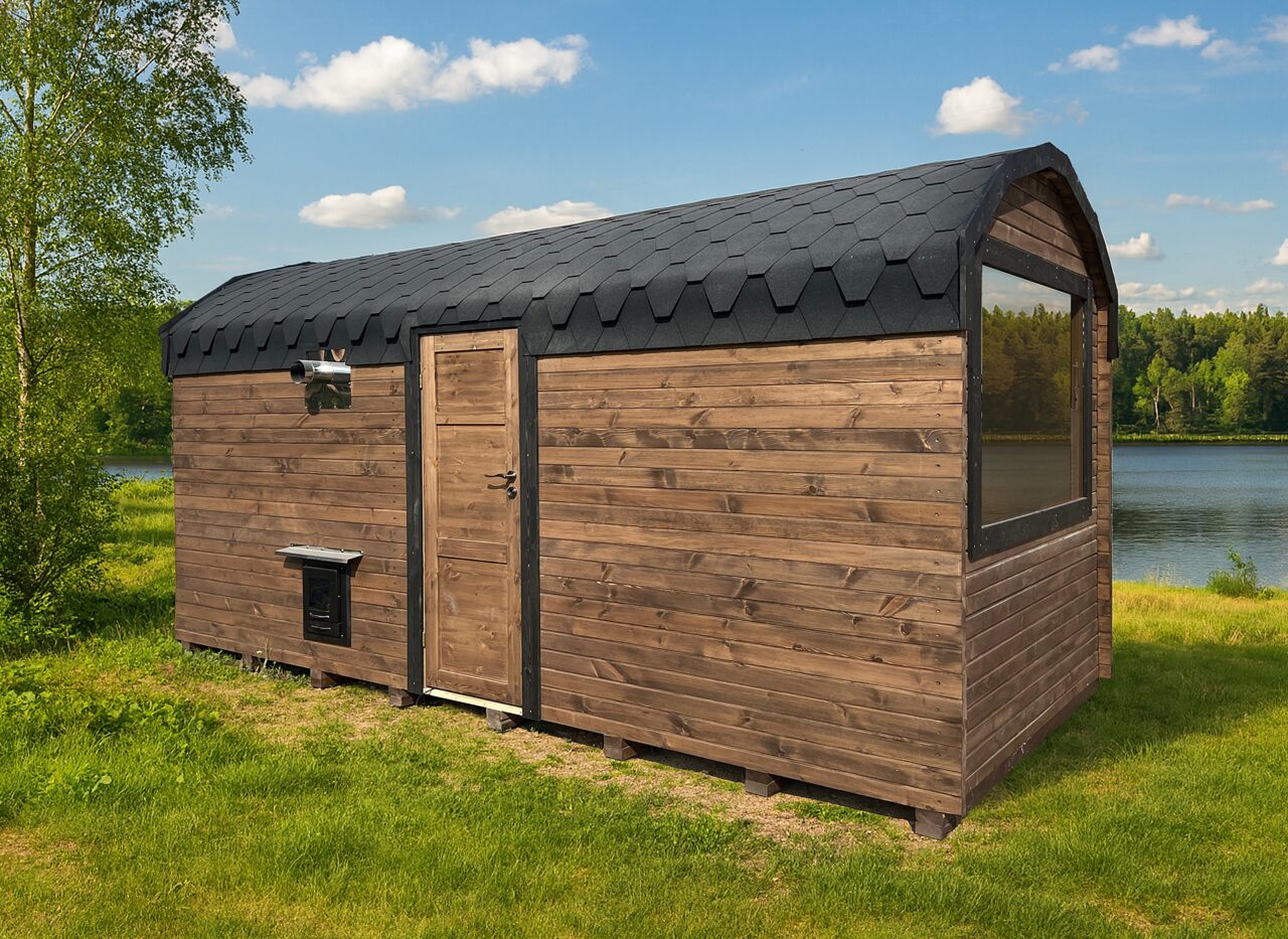 Feuerbad Sauna CERES auf einer Wiese, umgeben von Büschen und Bäumen. Im Hintergrund sieht man einen See und blauen Himmel