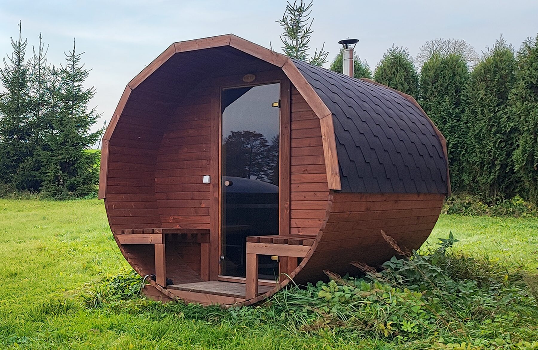Feuerbad Fass-Sauna JUNO auf einer Wiese mit Bäumen und blauem Himmel im Hintergrund