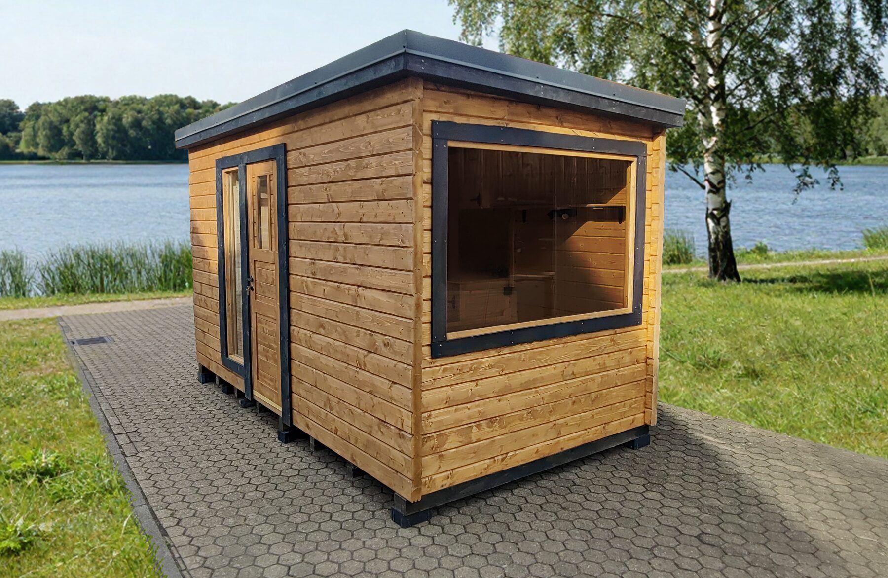 Exterior view of the Feuerbad tiny house DELUXE with wooden cladding and dark window frames, placed on paved ground by a lake, surrounded by green grass, trees, and a calm natural landscape.