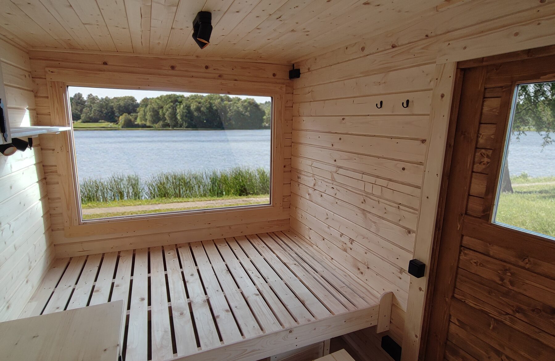 Interior view of the Feuerbad tiny house DELUXE featuring a light wooden interior, a spacious sleeping area, and a large panoramic window overlooking a lake and reeds, filled with natural light and a close-to-nature atmosphere.