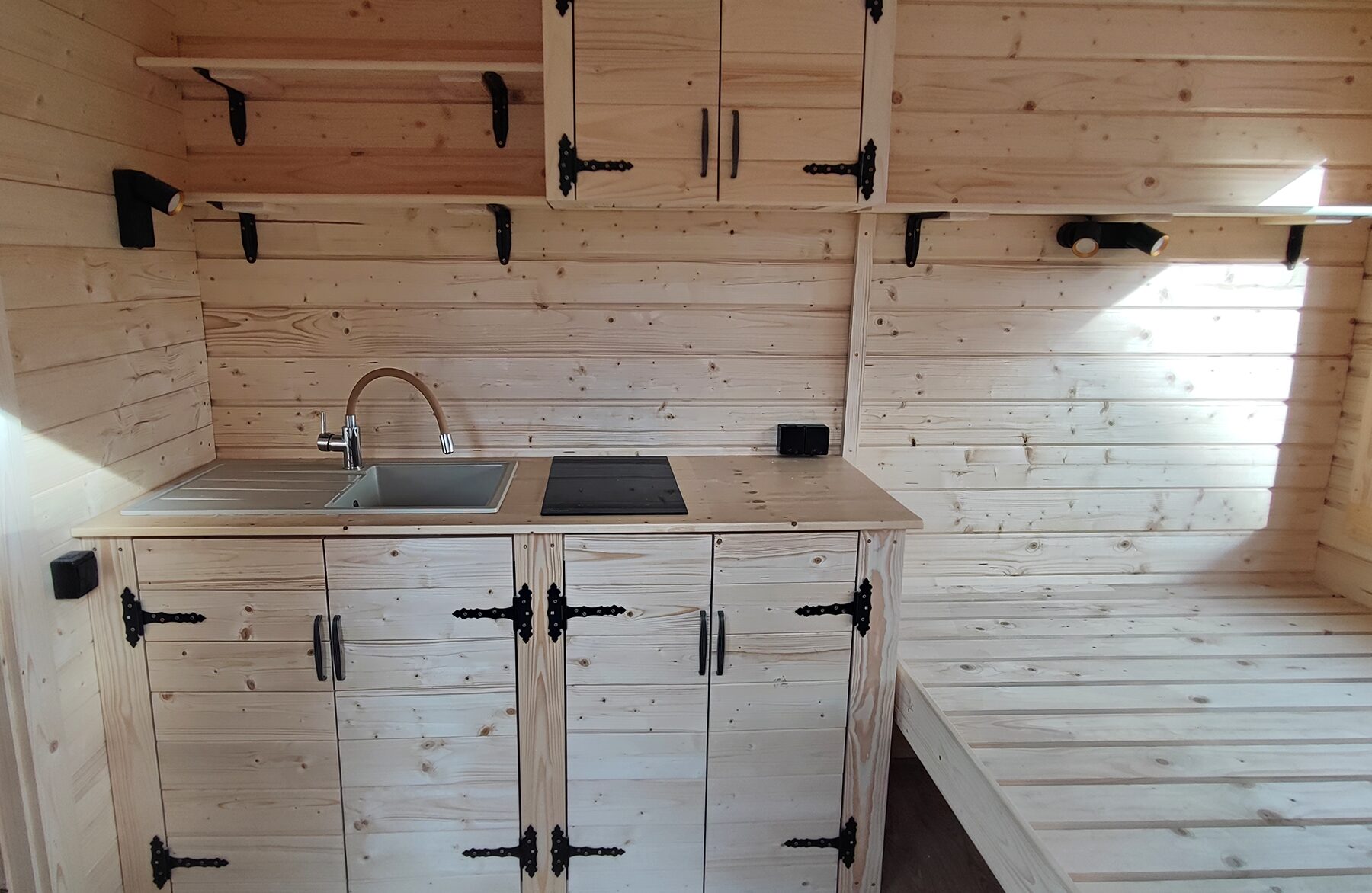 view of a Feuerbad tiny house DELUXE featuring a compact wooden kitchen with a sink, countertop with cooktop, base cabinets, and open shelves. The light wood paneling, black hardware, and integrated lighting create a functional and cozy living space.