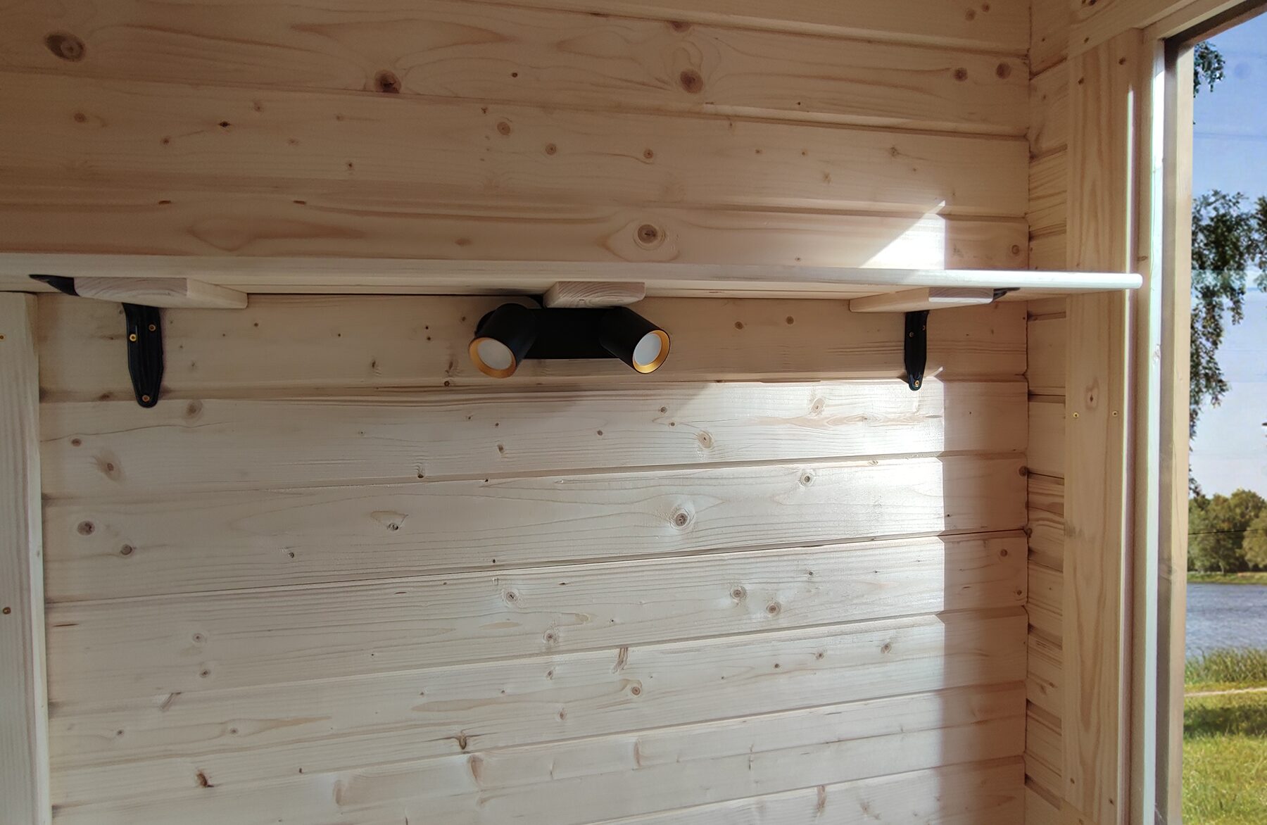 Detail view of the interior wall of a Feuerbad tiny house DELUXE featuring light wood paneling, a slim wall shelf mounted on black metal brackets, and an integrated black double wall light. Natural daylight enters from a side window, enhancing the warm and natural living atmosphere.