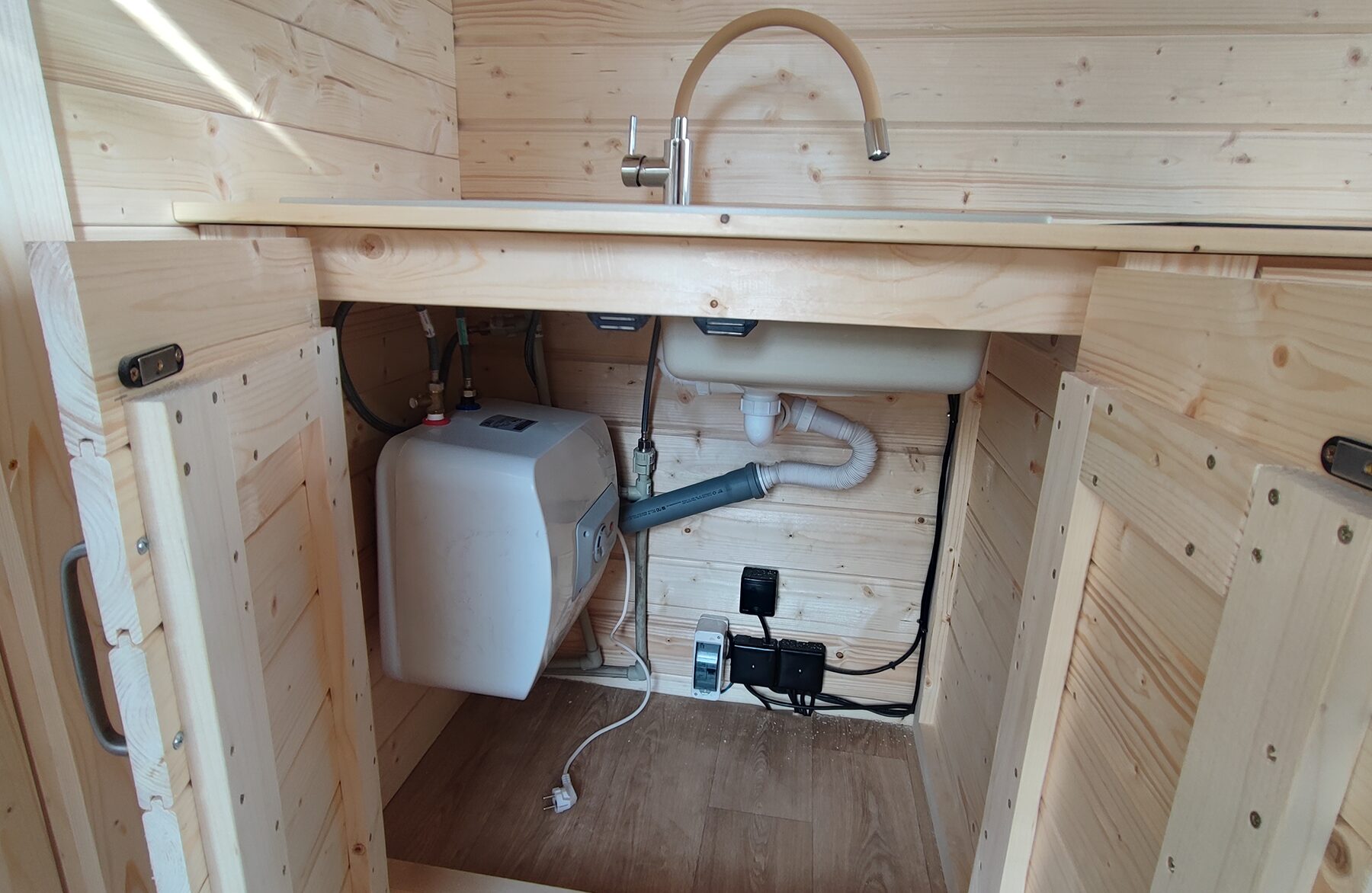 Detailed view of the kitchen area in a Feuerbad tiny house DELUXE with open base cabinet doors: visible are the sink with faucet, the water installation including a water heater, drainage pipes, and electrical connections. The light wood paneling highlights the functional and space-efficient design.