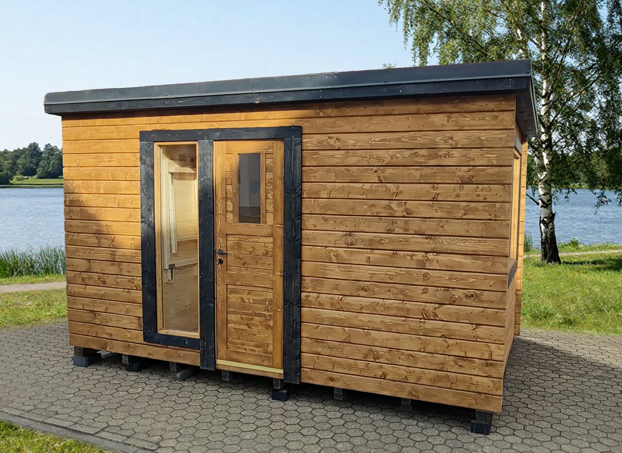Exterior view of the Feuerbad tiny house DELUXE with wooden cladding and dark window frames, placed on paved ground by a lake, surrounded by green grass, trees, and a calm natural landscape.