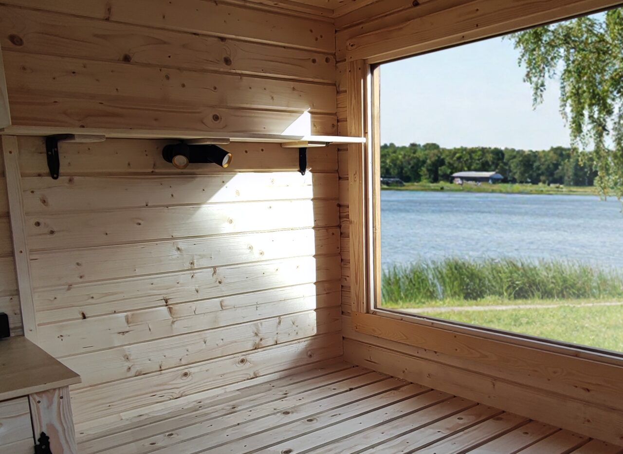 Interior view of the Feuerbad tiny house DELUXE featuring a light wooden interior, a spacious sleeping area, and a large panoramic window overlooking a lake and reeds, filled with natural light and a close-to-nature atmosphere.