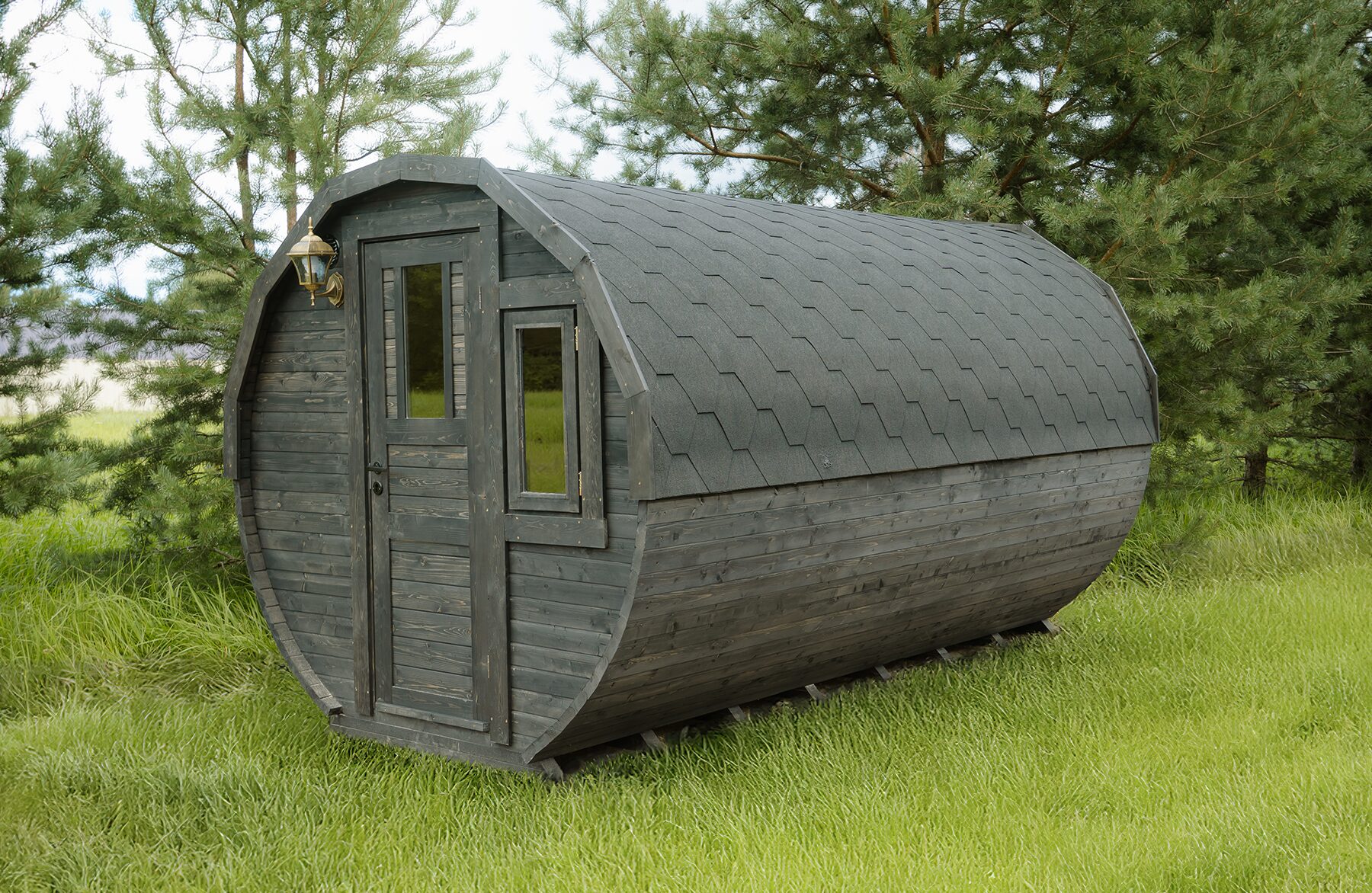 Außenansicht des Feuerbad Tiny House JUNO mit dunkler Holzverkleidung und gewölbter Form, ausgestattet mit Eingangstür, Seitenfenster und Dachschindeln, platziert auf einer grünen Wiese und umgeben von Bäumen