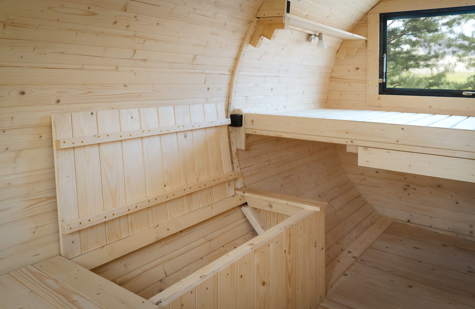 Detailansicht des Innenraums des Feuerbad Tiny House JUNO mit heller Holzverkleidung. Zu sehen sind ein erhöhter Liegebereich, eine aufklappbare Sitzbank sowie ein Fenster mit Blick ins Grüne. Die klare Holzstruktur und die durchdachte Konstruktion sorgen für eine helle und natürliche Atmosphäre