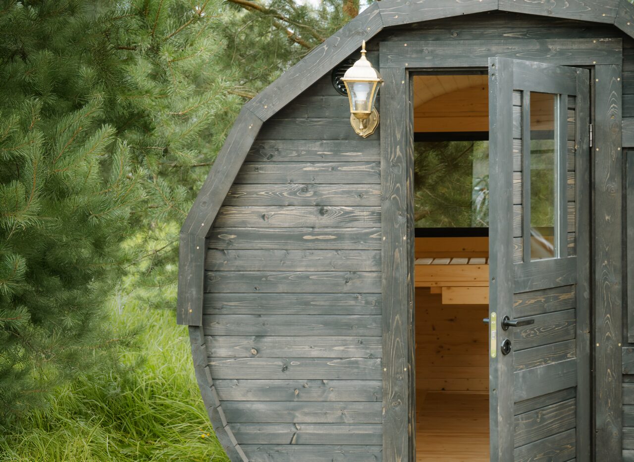 Detailansicht der Außenfront des Feuerbad Tiny House JUNO mit dunkel lasierter Holzverkleidung. Die geöffnete Tür gibt den Blick auf den hellen Innenraum mit Holzbänken frei. Eine klassische Außenlampe ist an der Fassade montiert, das Tiny House steht eingebettet in grünes Gras und umgeben von Nadelbäumen
